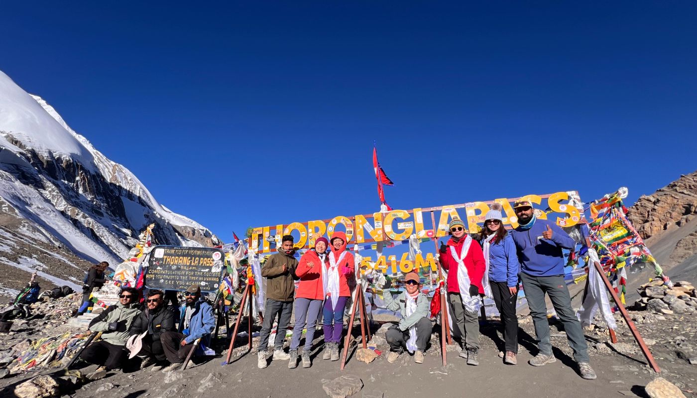 Trekkers celebrating at Thorong La Pass (5,416 m), the highest point of the Annapurna Circuit Trek, with snow-covered peaks and prayer flags under a clear blue Himalayan sky.